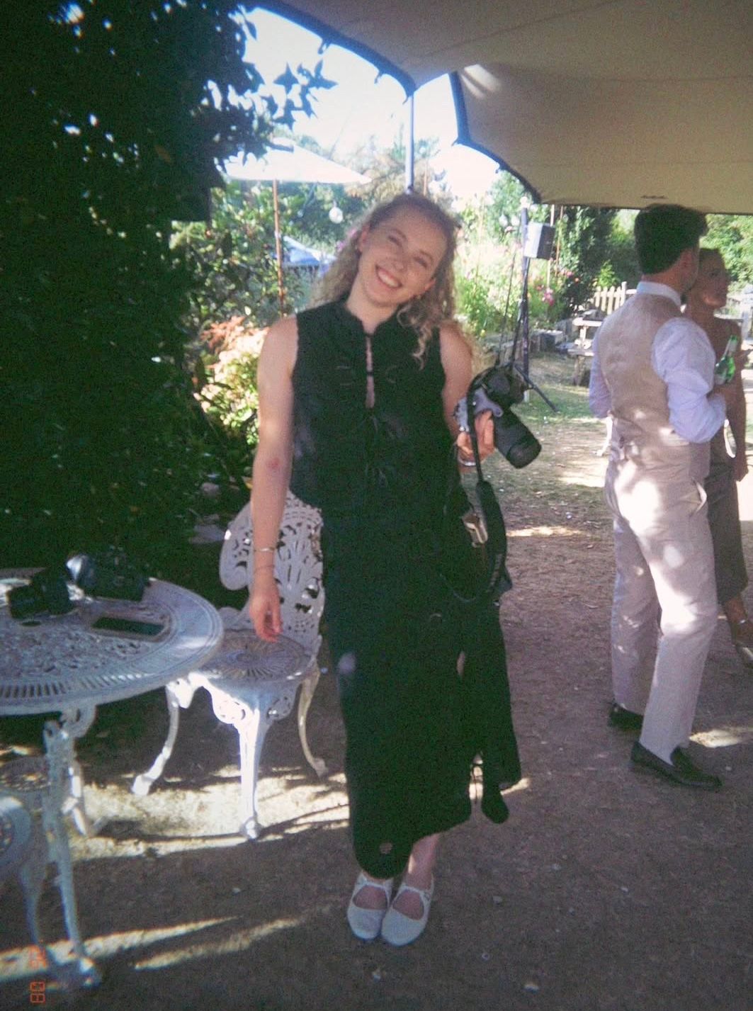 Woman in black dress holding camera smiles outdoors at garden party under blue umbrella.