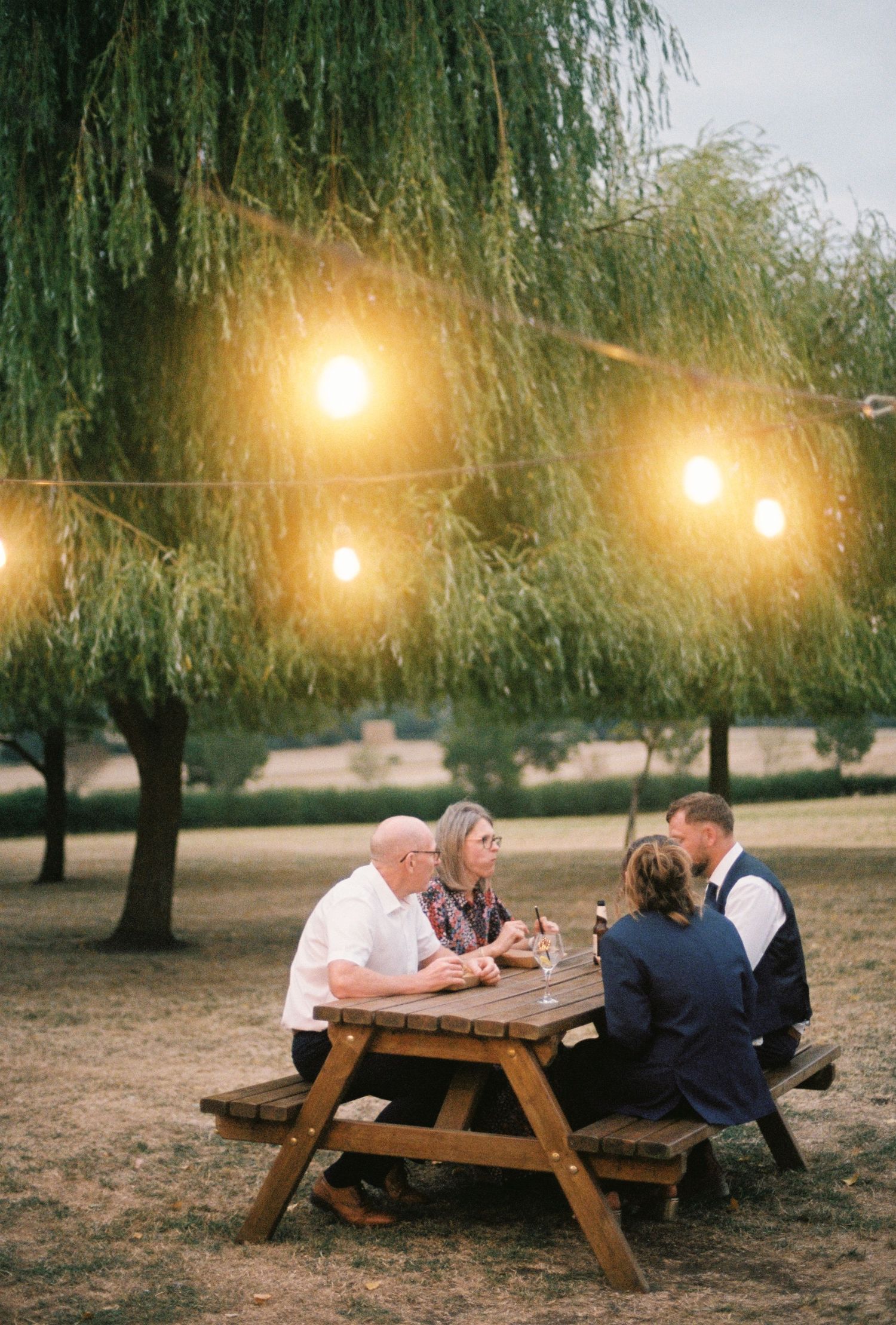 Four guests sit at a wooden picnic table under glowing string lights and a weeping willow at a twilight outdoor event.
