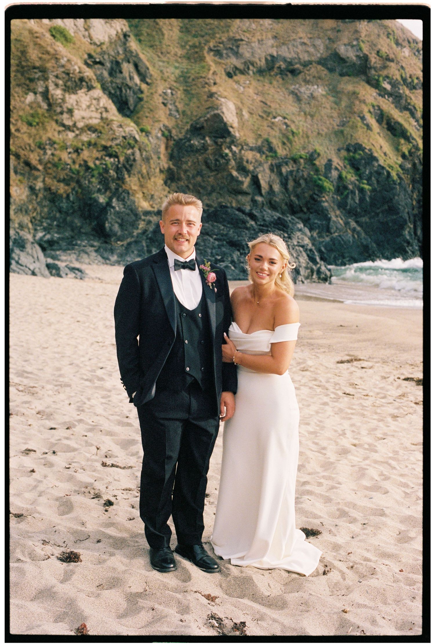 Bride and groom posing on sandy beach with dramatic cliffs, bride in off-shoulder white gown, groom in navy tuxedo.