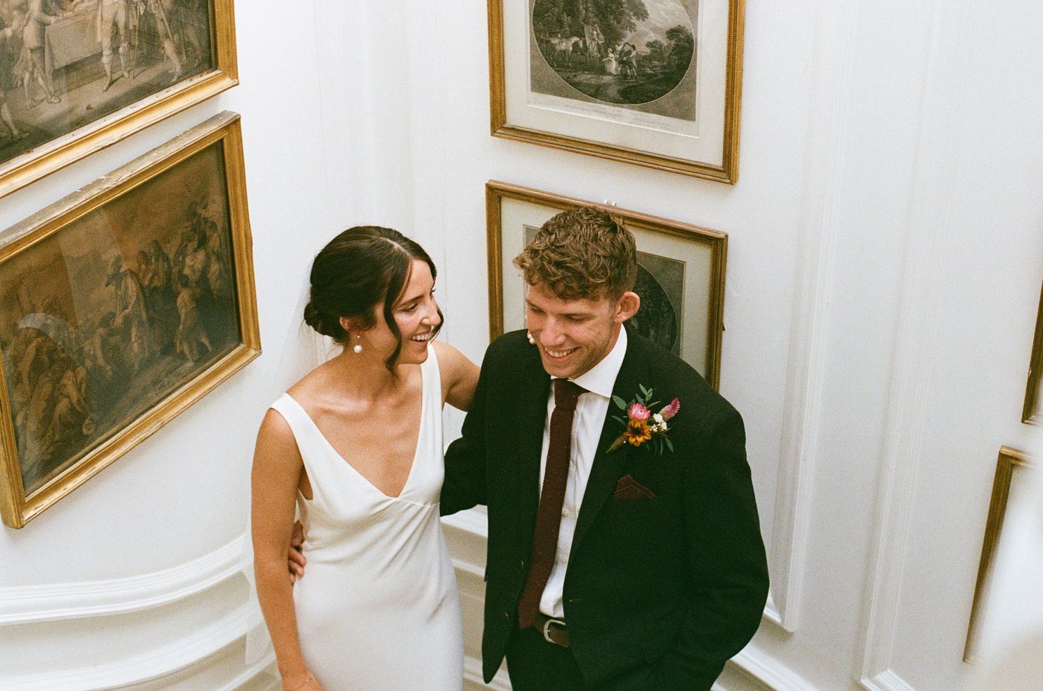 Happy bride and groom laughing together on a staircase surrounded by ornate gold-framed paintings.