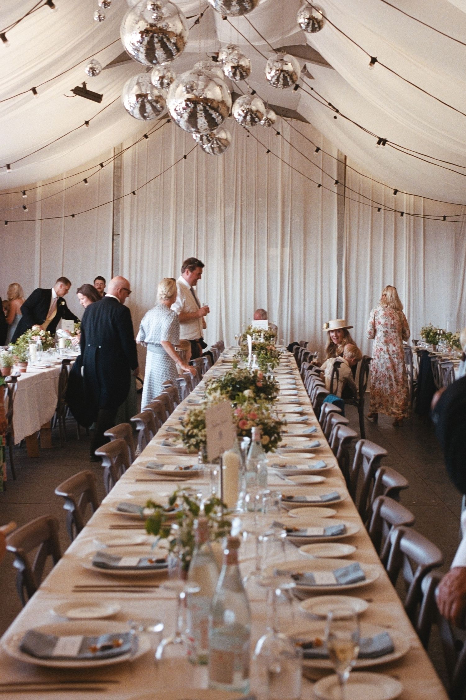 Elegant wedding reception in a draped marquee tent with long tables, floral centerpieces, and silver disco ball chandeliers.