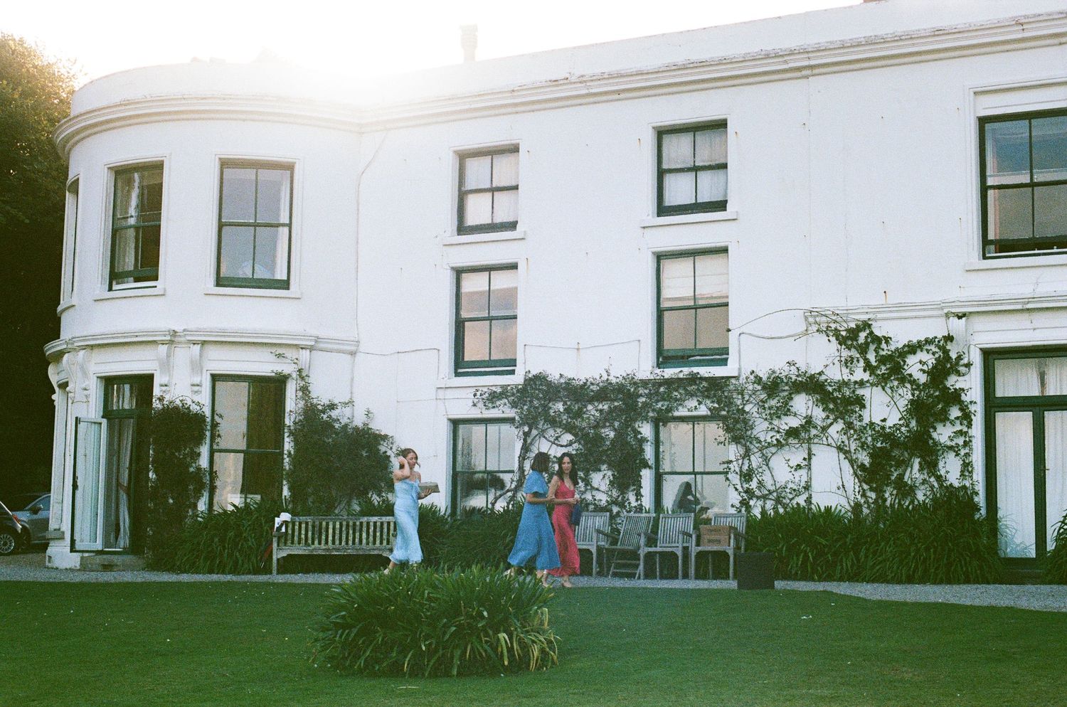 Three guests pose on the lawn of a white country house venue at golden hour during a wedding.