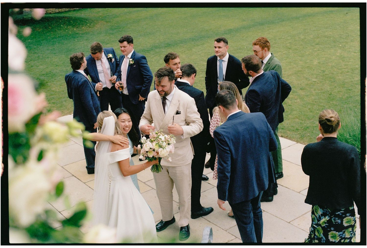 Bride and groom in elegant attire surrounded by groomsmen in navy suits at an outdoor wedding reception.