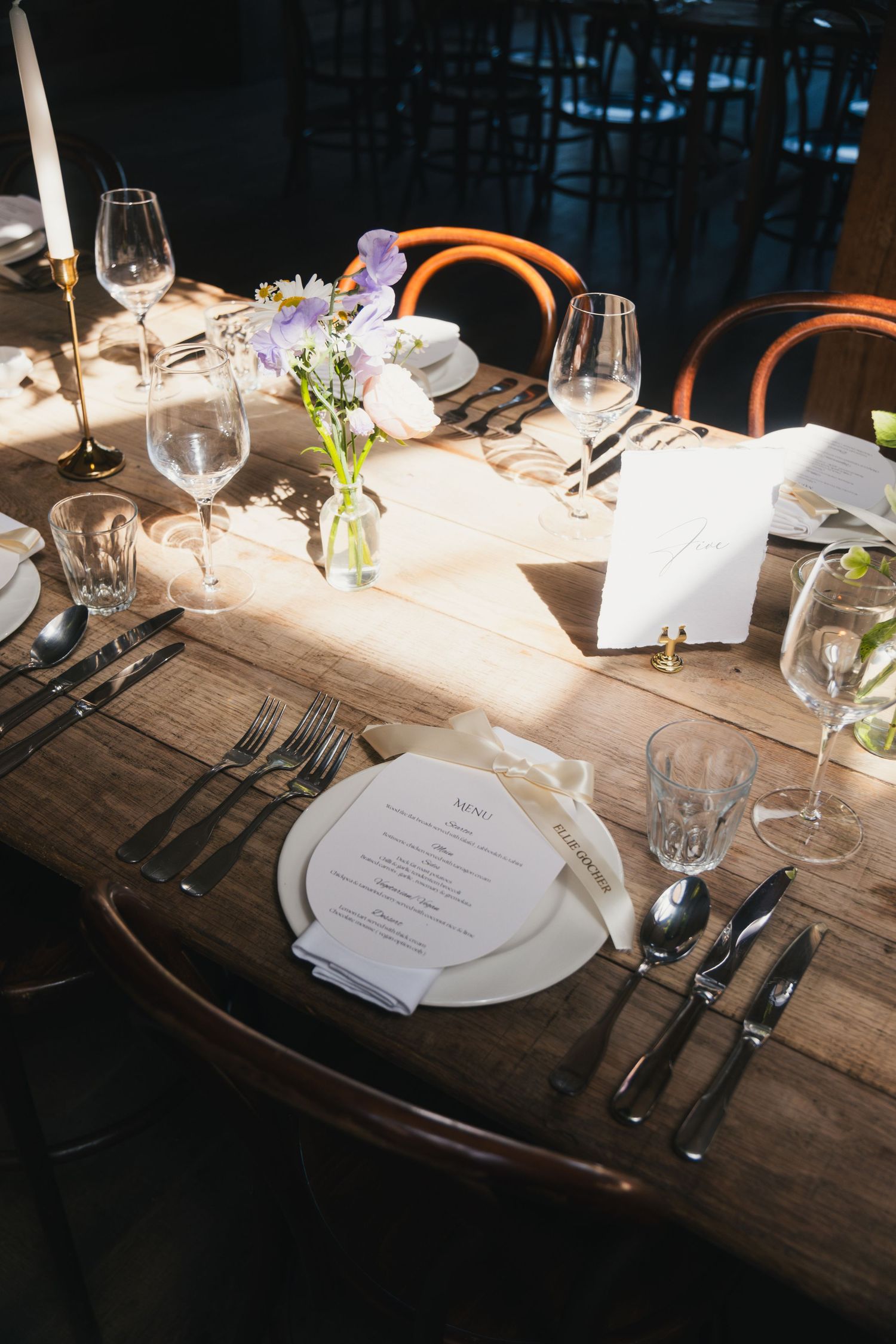 Elegant rustic wooden table set with white plates, menus, wine glasses, candles, and wildflower centerpieces for a formal event.