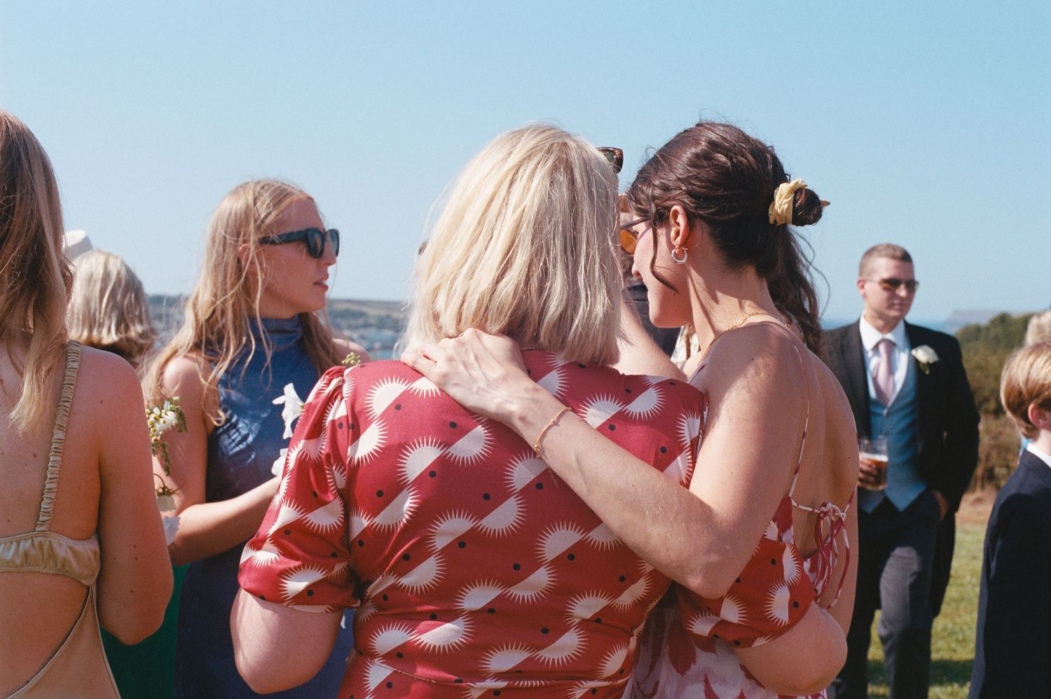 Two women hugging at an outdoor wedding reception on a sunny day with guests and ocean views in the background.