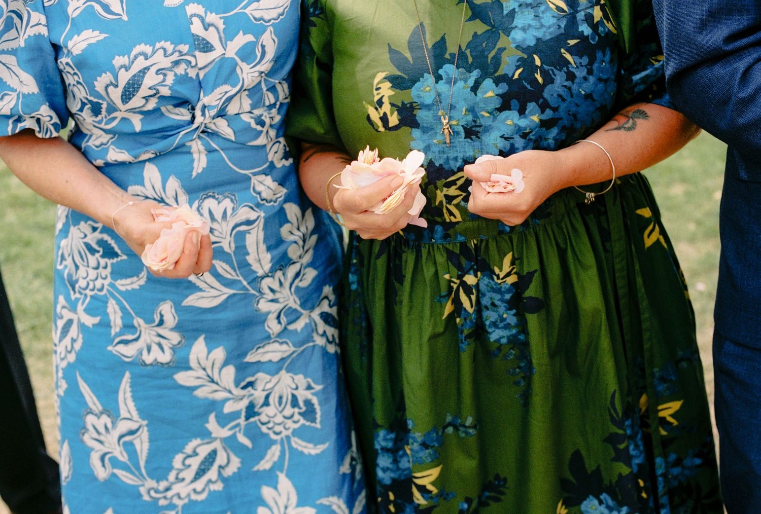 Three women in floral dresses holding white flowers, showcasing blue and green botanical prints at an outdoor event.
