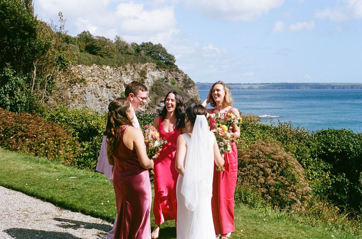 Bride in white gown with bridesmaids in pink dresses laughing together on a coastal garden path.