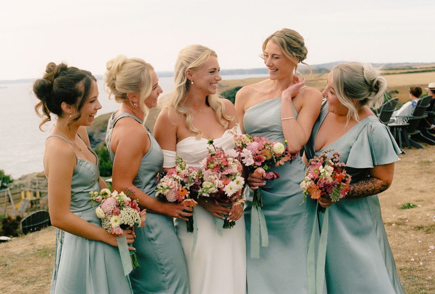 Bride in white strapless gown with bridesmaids in sage green dresses holding pink bouquets at outdoor coastal wedding.