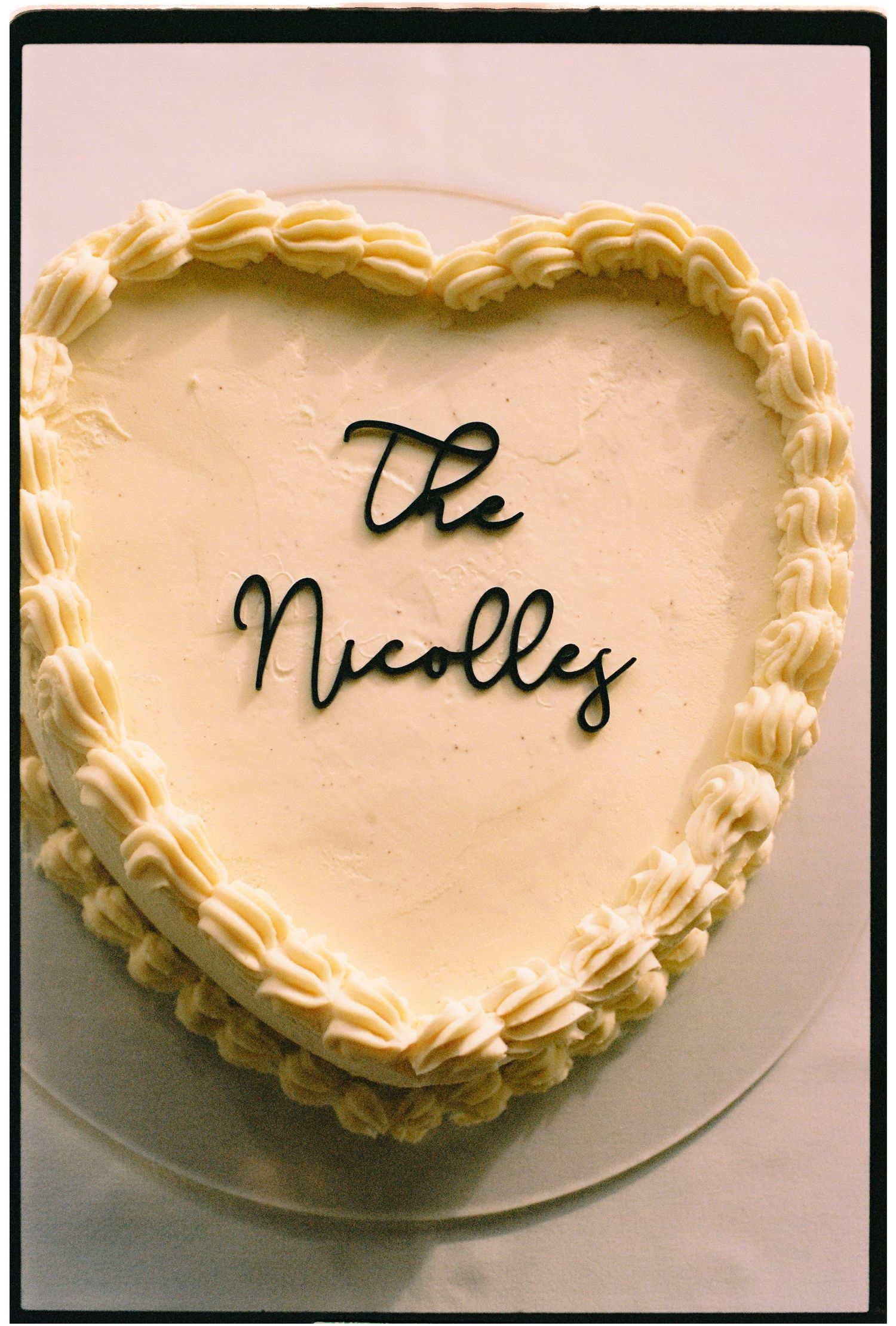 Heart-shaped white frosted cake with piped borders and black script topper reading The Nicolles on a glass stand.