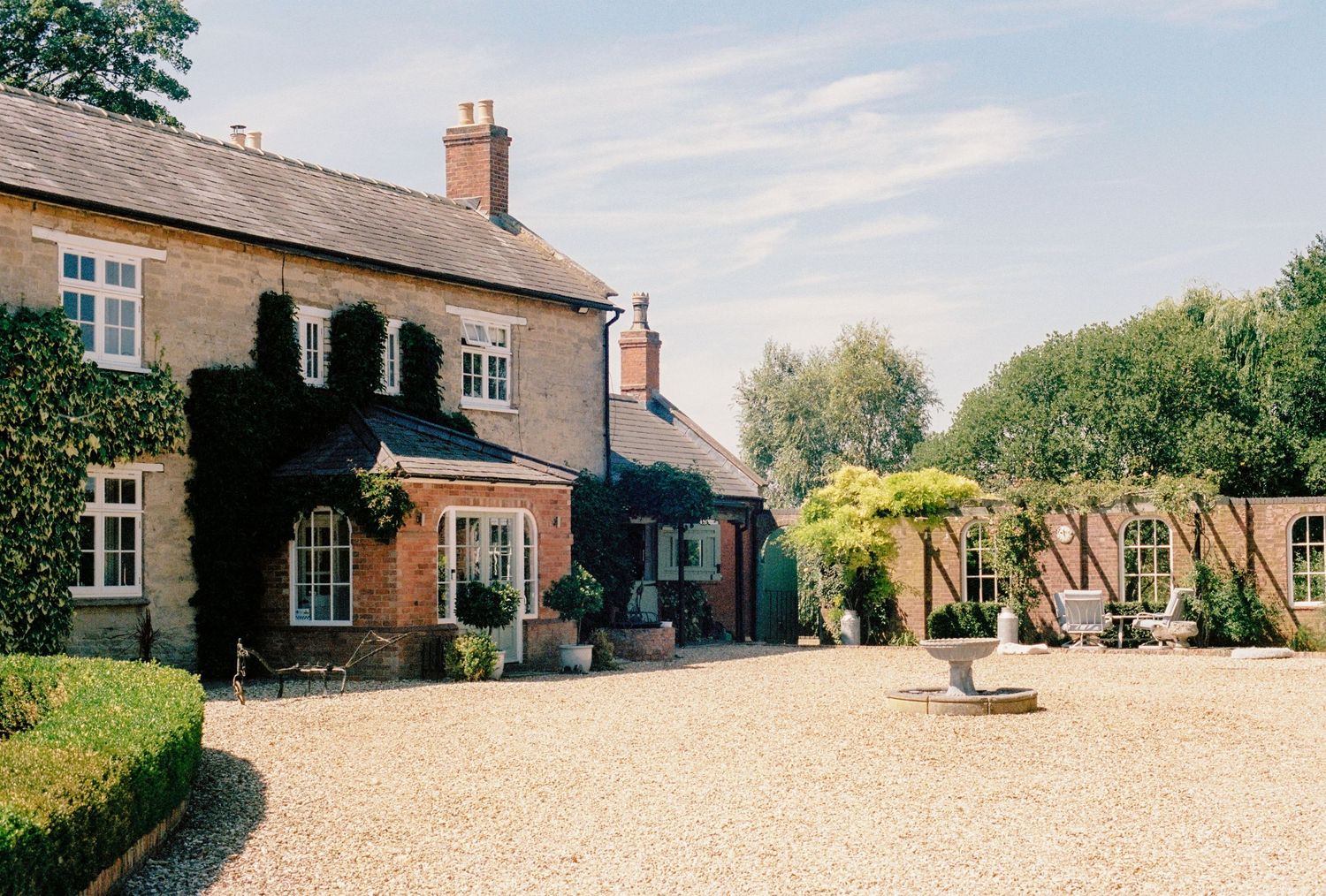 Charming ivy-covered English countryside manor house with gravel courtyard, manicured hedges, and sunny blue sky.