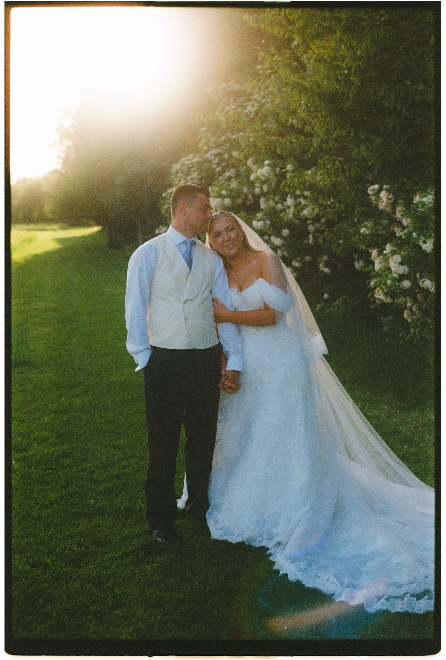 Bride and groom share a romantic golden hour moment outdoors, bride in lace gown with veil, groom in white vest.