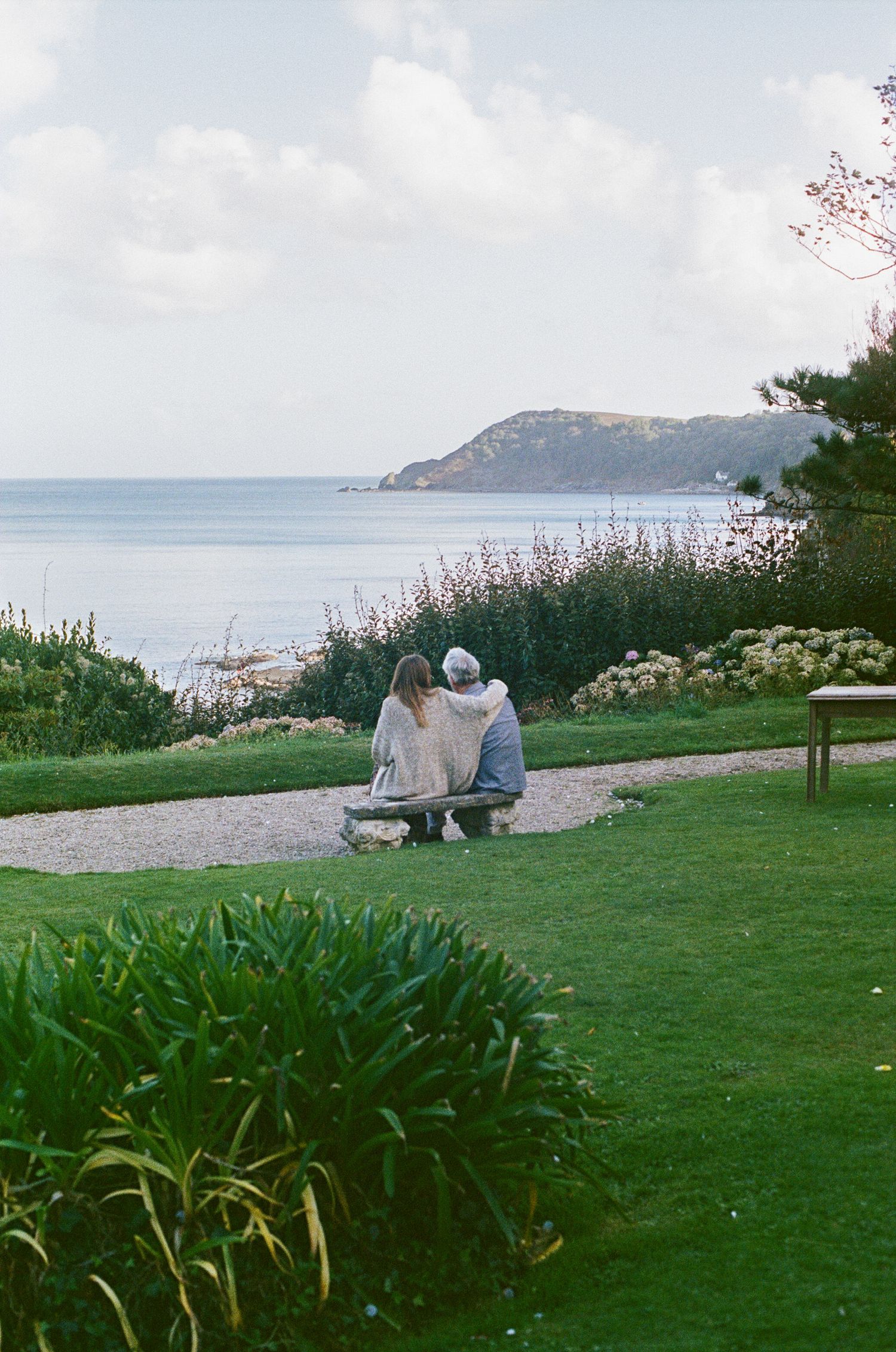 Two people sit on a bench overlooking a scenic coastal view with green hills and calm ocean waters.