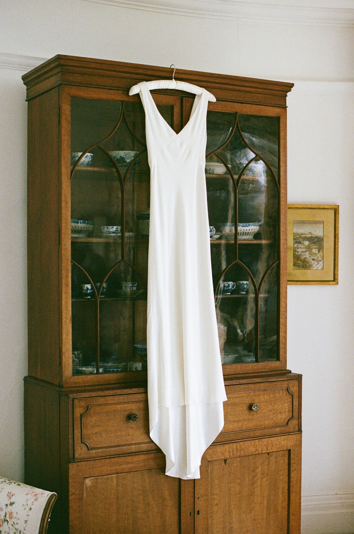 White wedding dress hanging on a wooden antique china cabinet in a vintage-styled room.