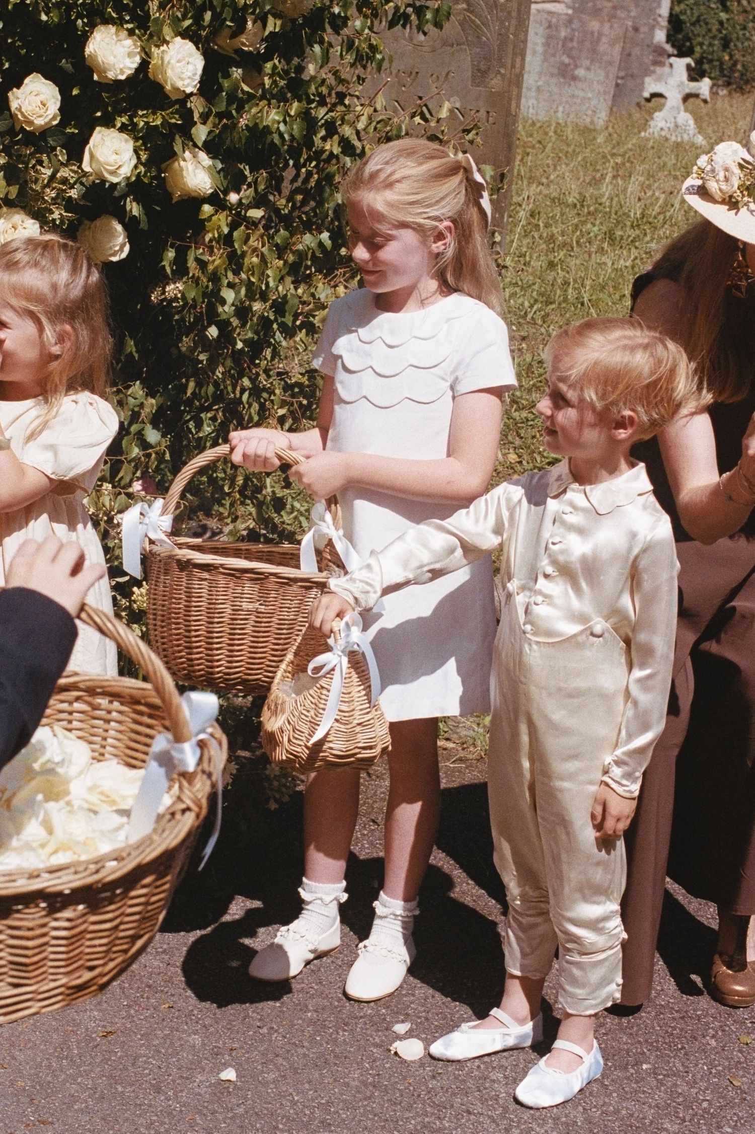 Children in formal wedding attire hold wicker baskets outdoors at a sunny garden wedding ceremony.