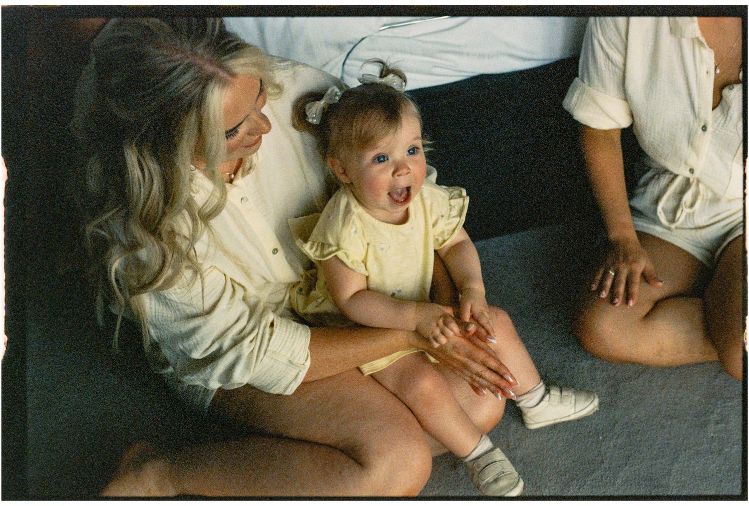 A blonde woman smiles at a laughing baby girl in a yellow dress sitting on the floor together.