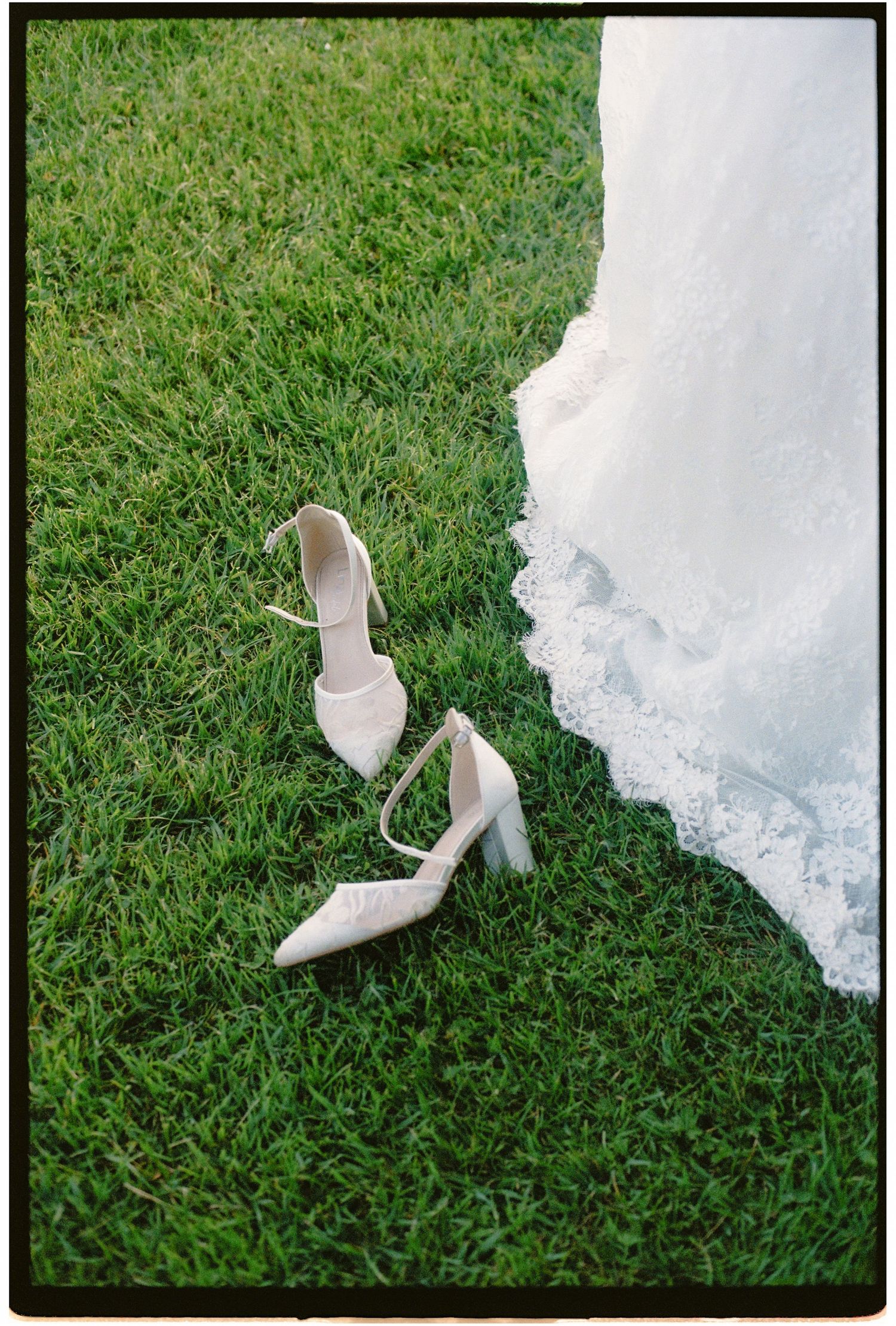 Elegant white bridal heels with ankle straps resting on lush green grass beside a lace wedding gown hem.