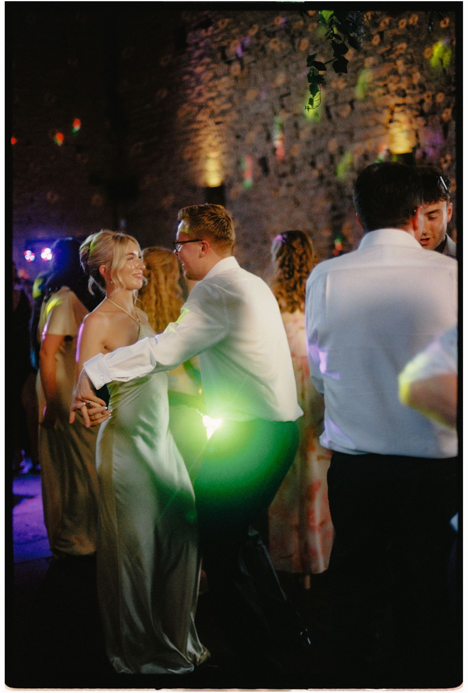 Bride and groom dancing joyfully at a wedding reception in a rustic stone venue with colorful lights.
