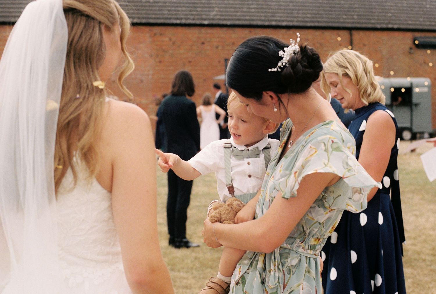 Bride in white gown and veil greeting guests including woman holding young child in suit at outdoor wedding reception.