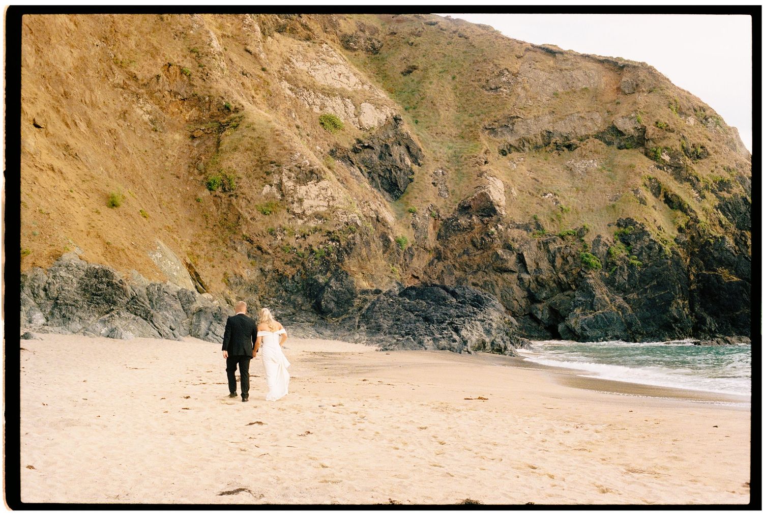A bride and groom walk hand in hand along a sandy beach with dramatic rocky cliffs in the background.