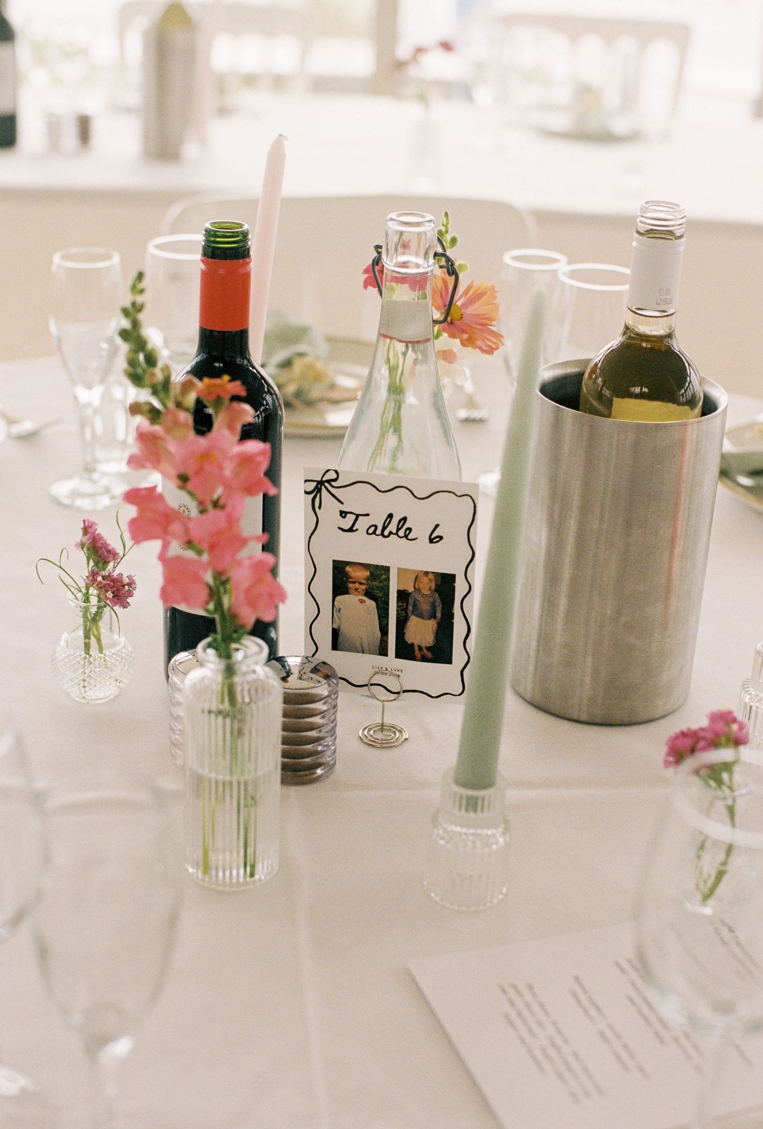 Wedding reception Table 6 centerpiece with red and white wine bottles, pink flowers in bud vases, and candles on white tablecloth.