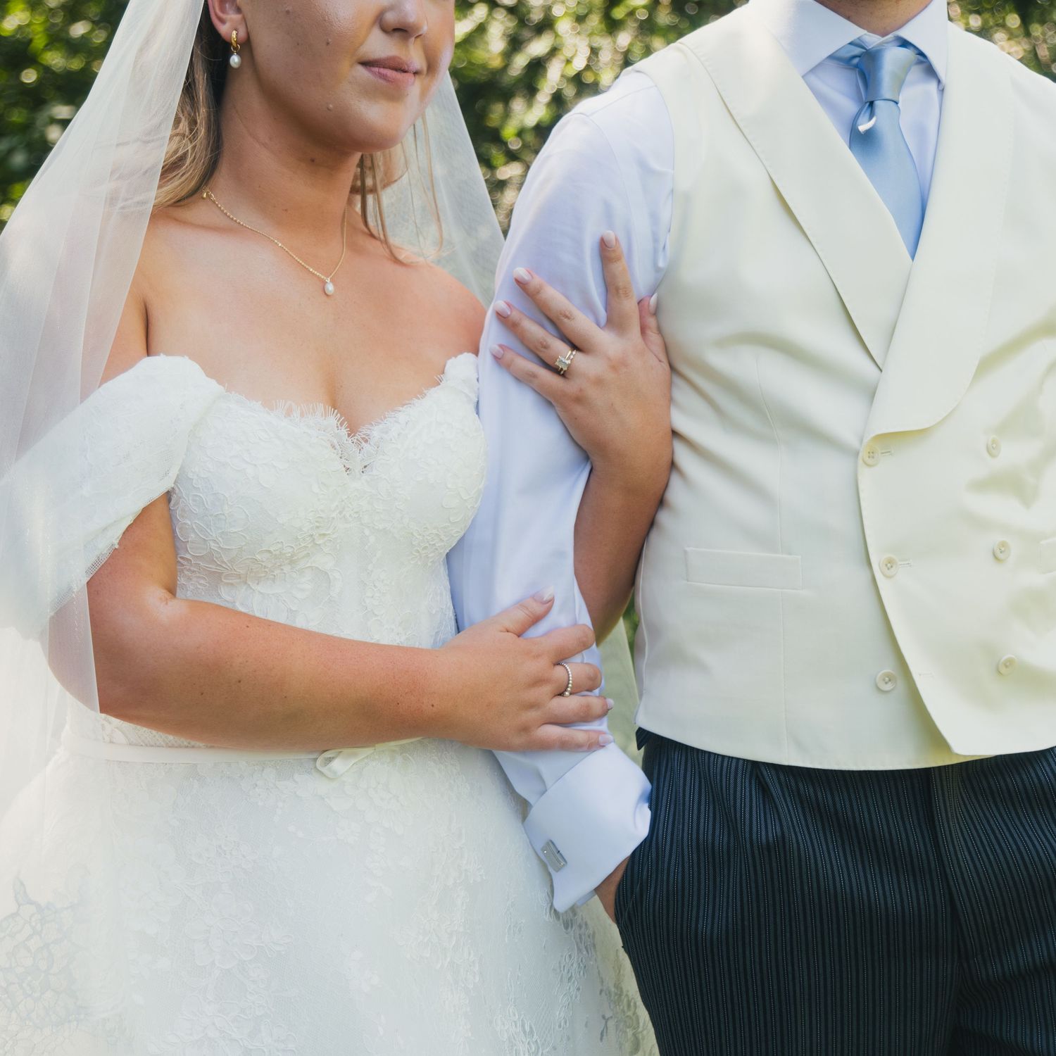 Bride in lace wedding gown and veil stands beside groom in cream suit, posing together outdoors on their wedding day.