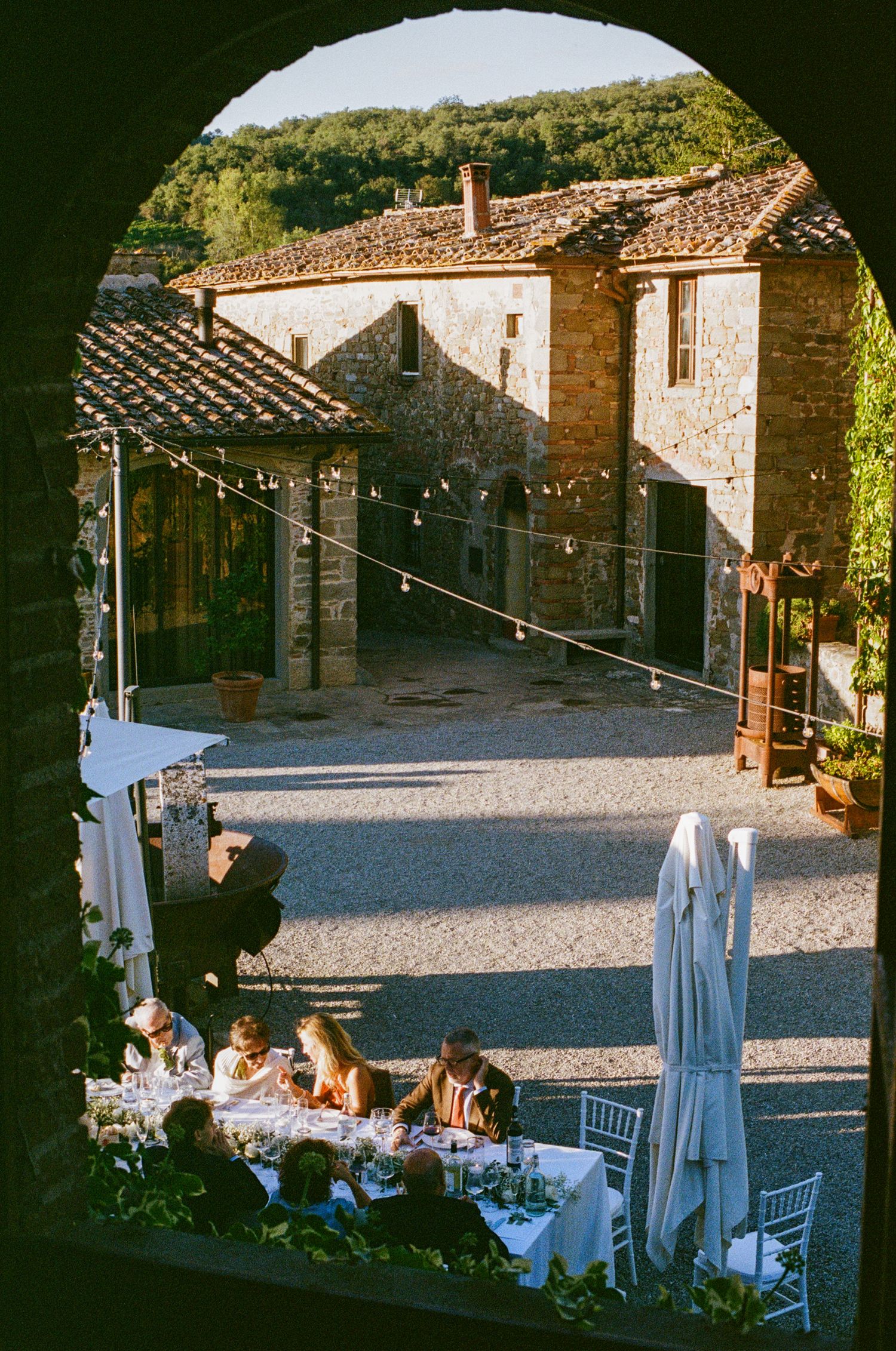 Wedding reception dinner viewed through stone arch at charming rustic Tuscan courtyard with string lights overhead.