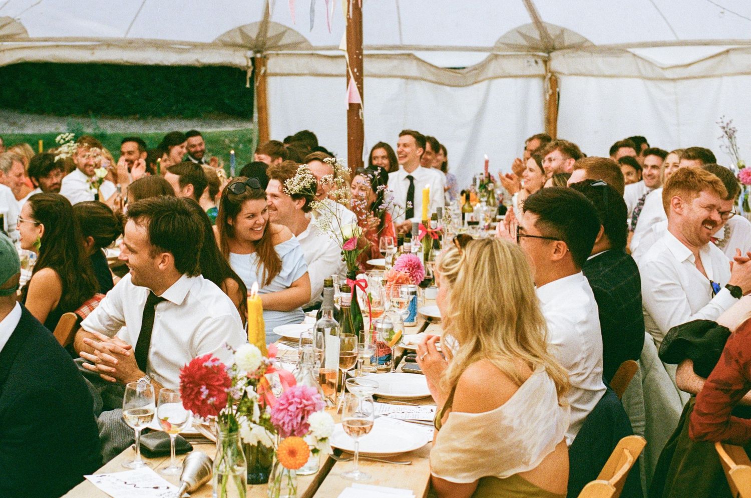 Large wedding reception crowd seated at long tables under a white marquee tent with colorful floral centerpieces.