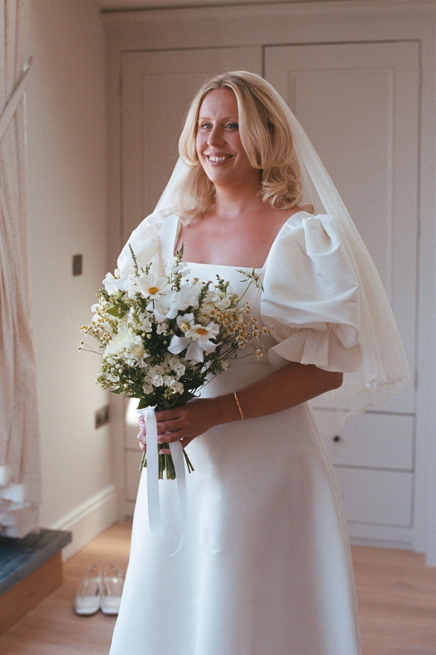 Blonde bride in a white puff-sleeve gown holding a wildflower bouquet, wearing a veil, smiling indoors.