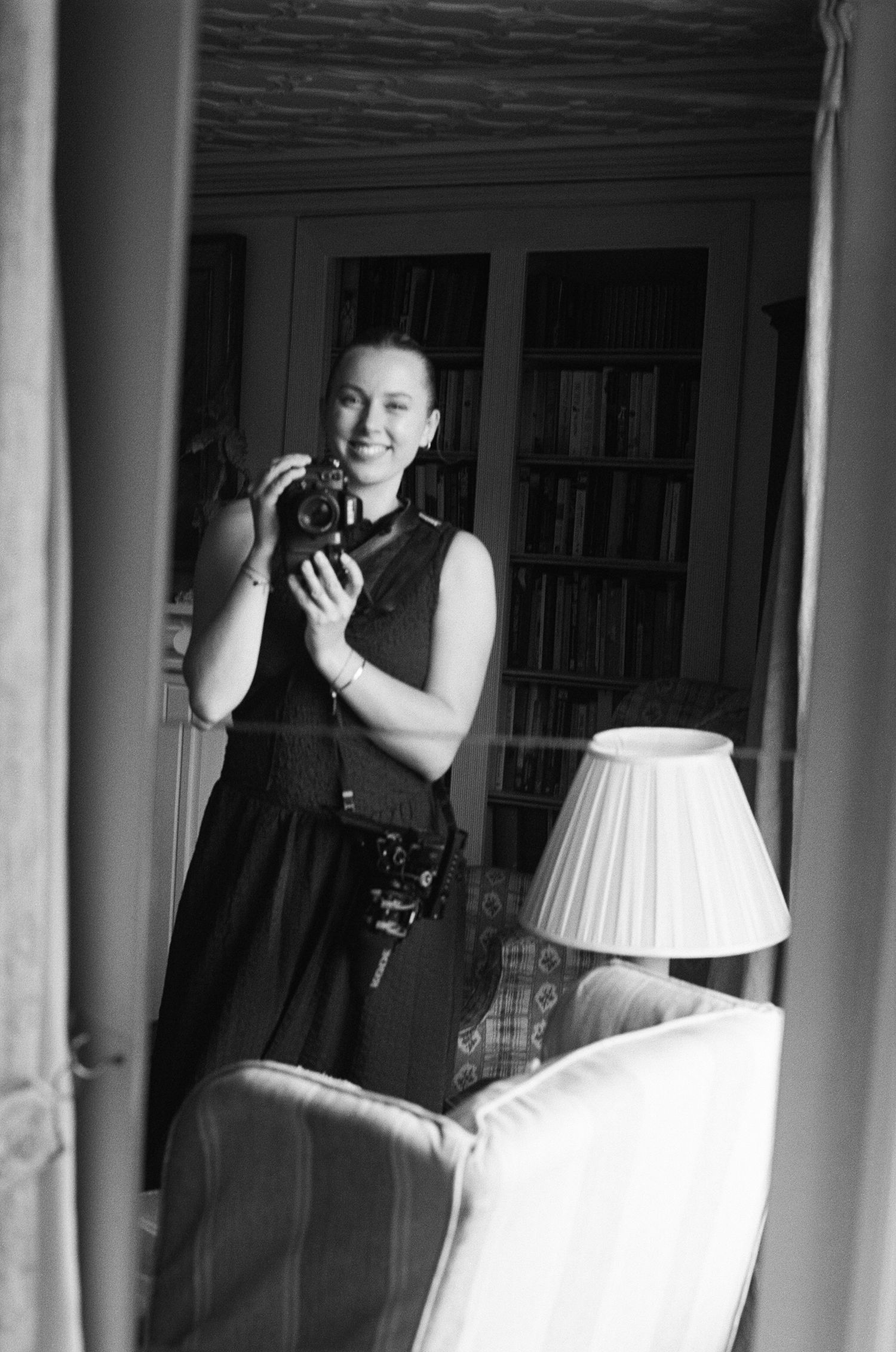 Black and white mirror selfie of a smiling woman in a sleeveless dress holding a camera in an elegant room.