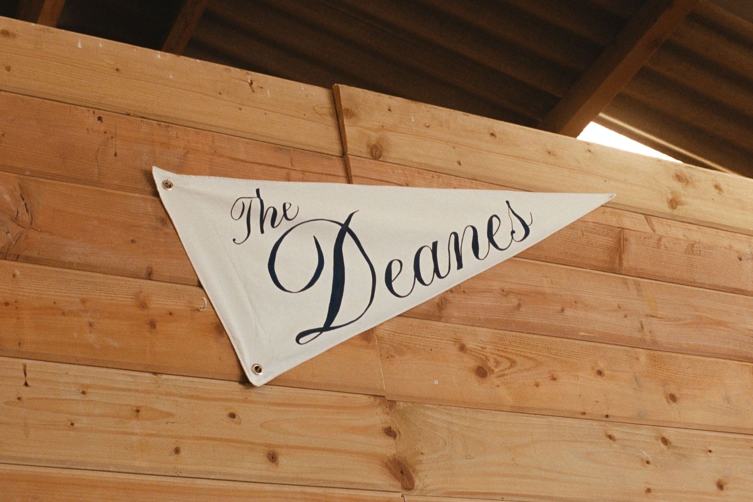 Triangular pennant banner reading The Deanes hanging on a wooden wall at a rustic wedding venue.