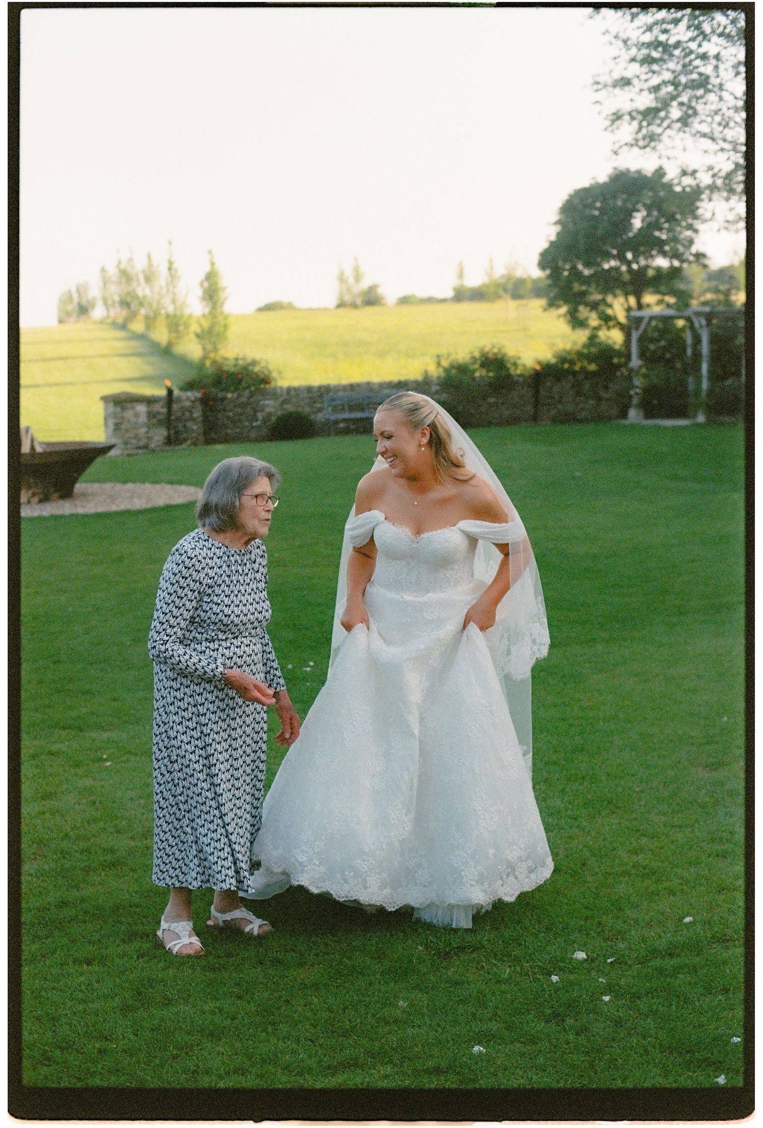 Bride in white off-shoulder gown with veil stands with elderly woman on green lawn at outdoor wedding venue.