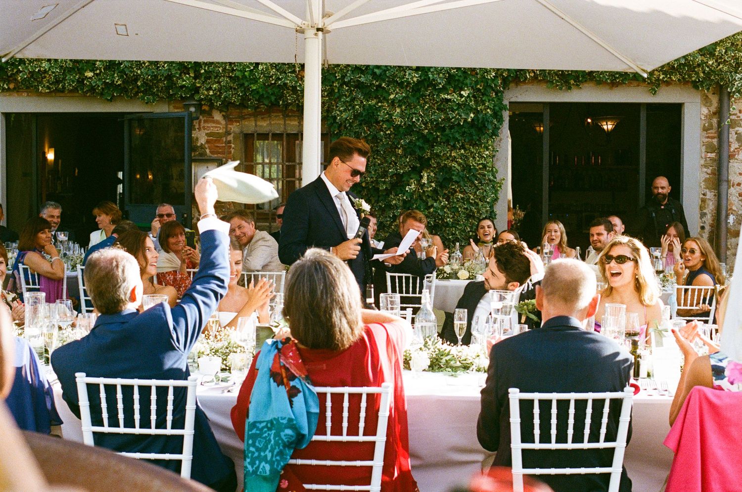Wedding guest raises hand during lively outdoor reception speech at rustic Tuscan venue with string lights.