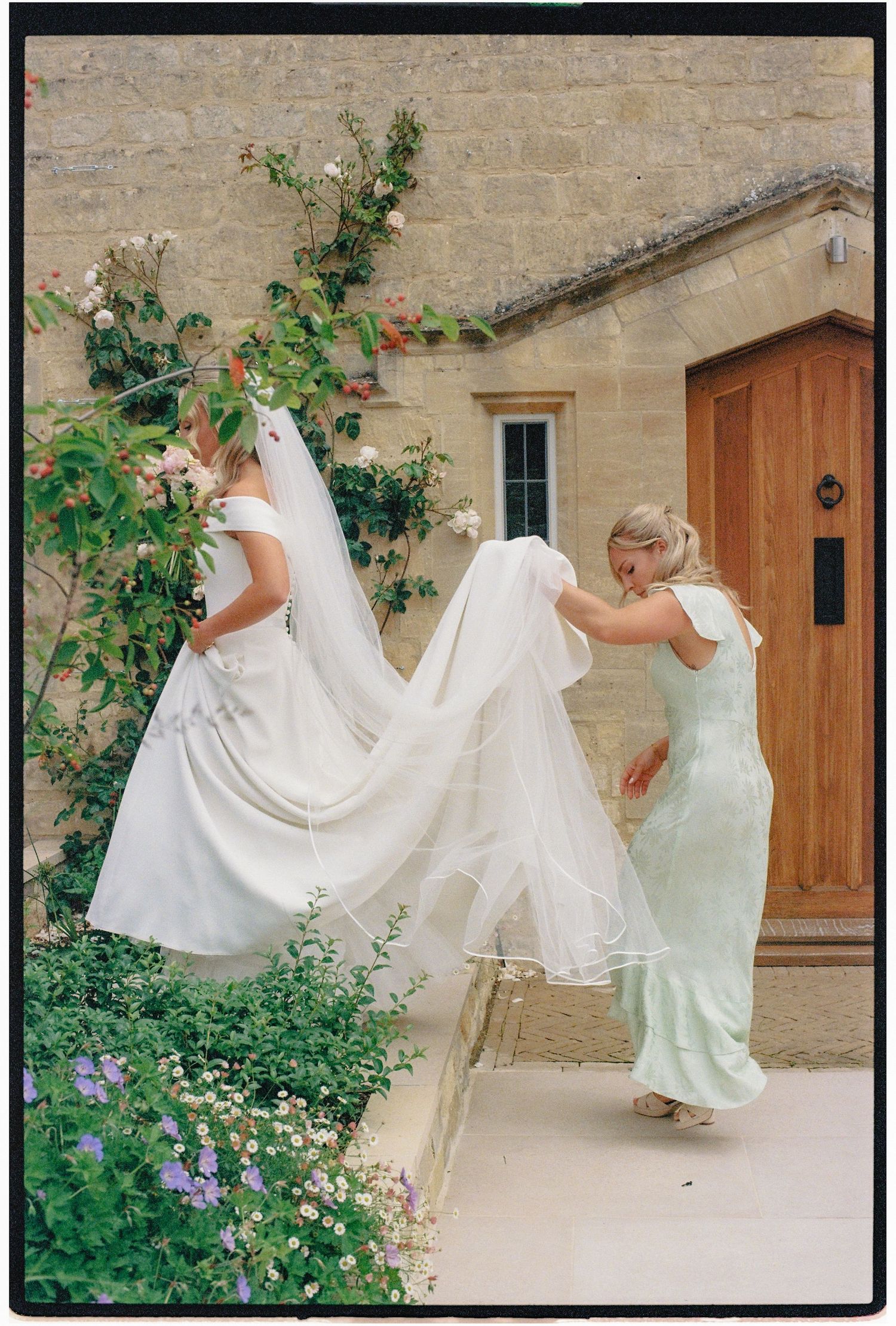 A bridesmaid in mint green adjusts the veil of a bride in white outside a stone building with climbing roses.