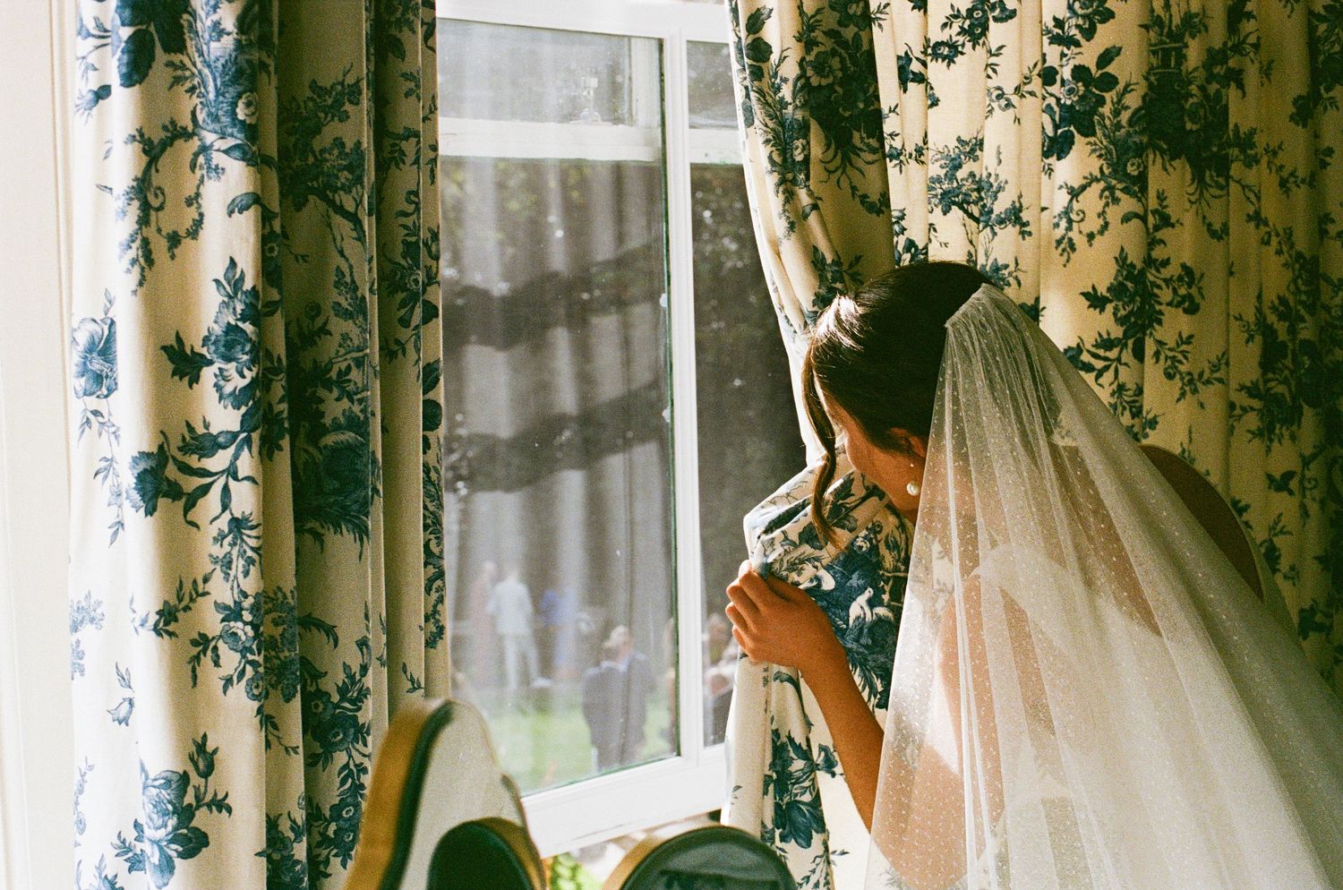 Bride wearing a white veil gazes out a window framed by blue and white floral curtains.