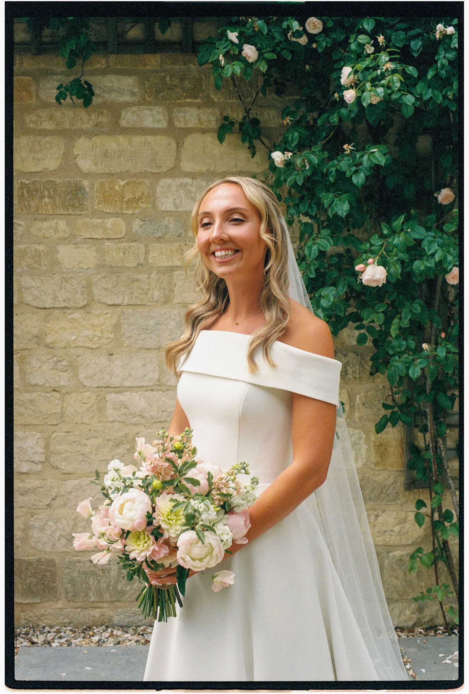 Smiling bride in off-shoulder white gown holding pink bouquet, standing before stone wall with climbing roses.