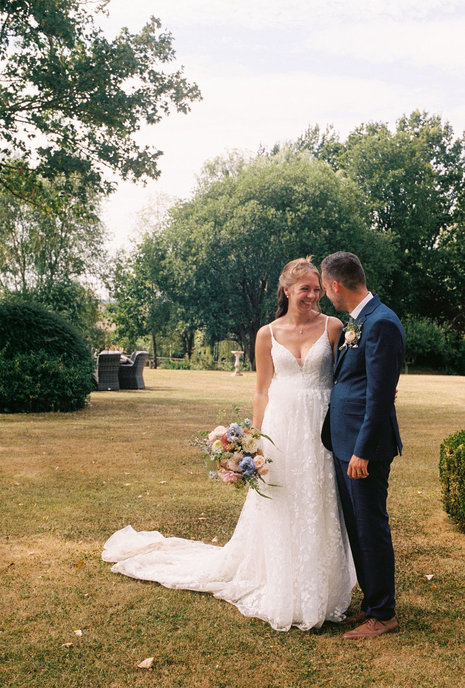 Bride in lace gown holding colorful bouquet stands with groom in navy suit in a lush garden setting.