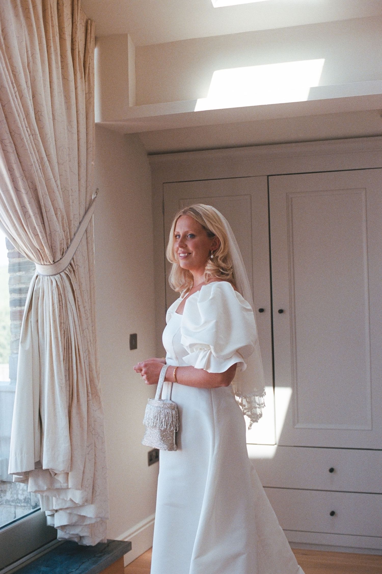 Bride in white puff-sleeve gown with veil and beaded clutch stands in elegant room with curtains.