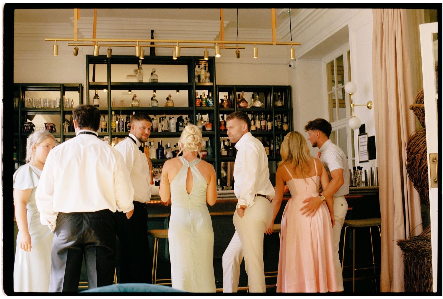 Elegant guests socializing at a stylish bar during a formal event, dressed in white, cream, and pink attire.