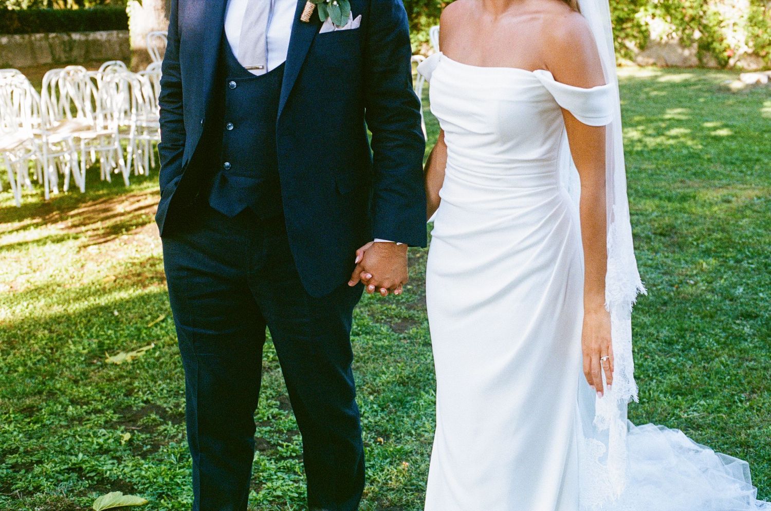 Bride in off-shoulder white gown and groom in navy suit hold hands at an outdoor garden wedding.
