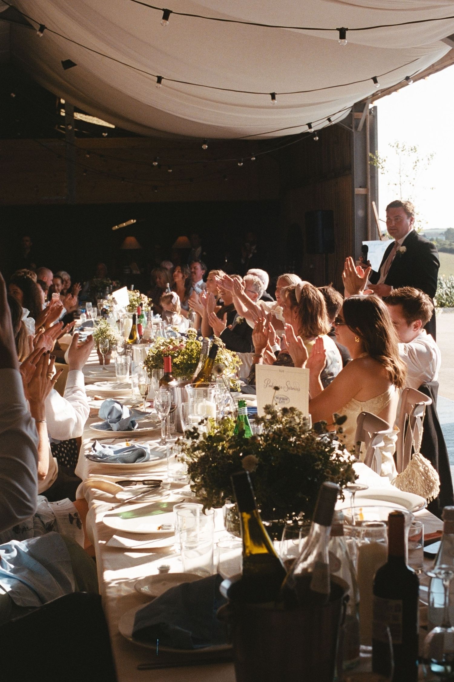 Guests raise glasses in a toast at an elegant wedding reception with long tables, floral centerpieces, and string lights.