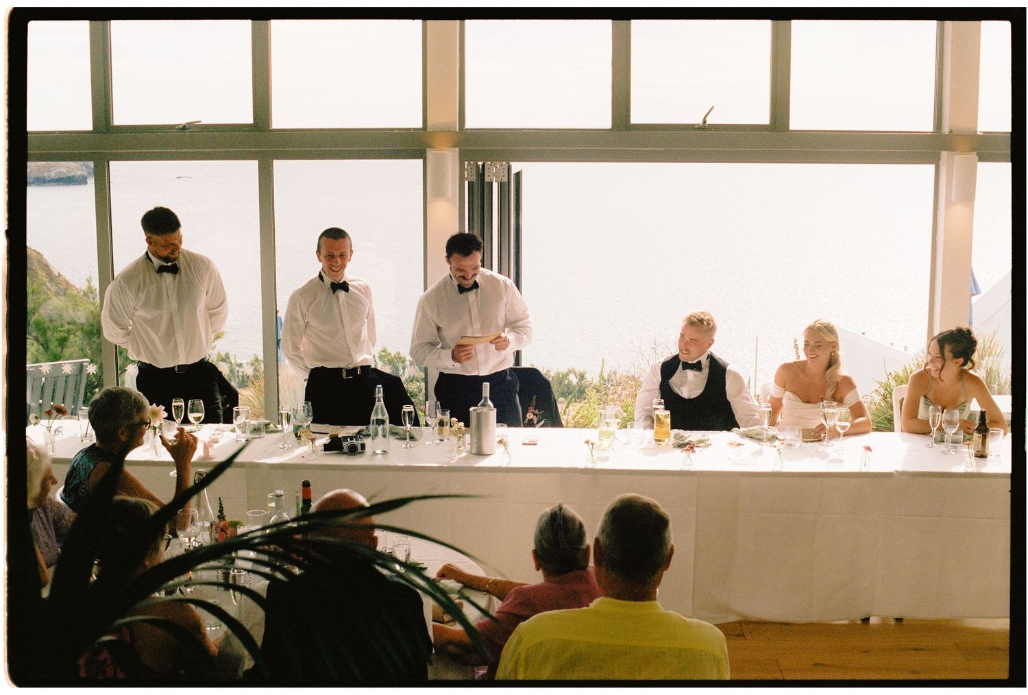 Wedding party stands at head table during reception speeches, with guests seated before large windows.