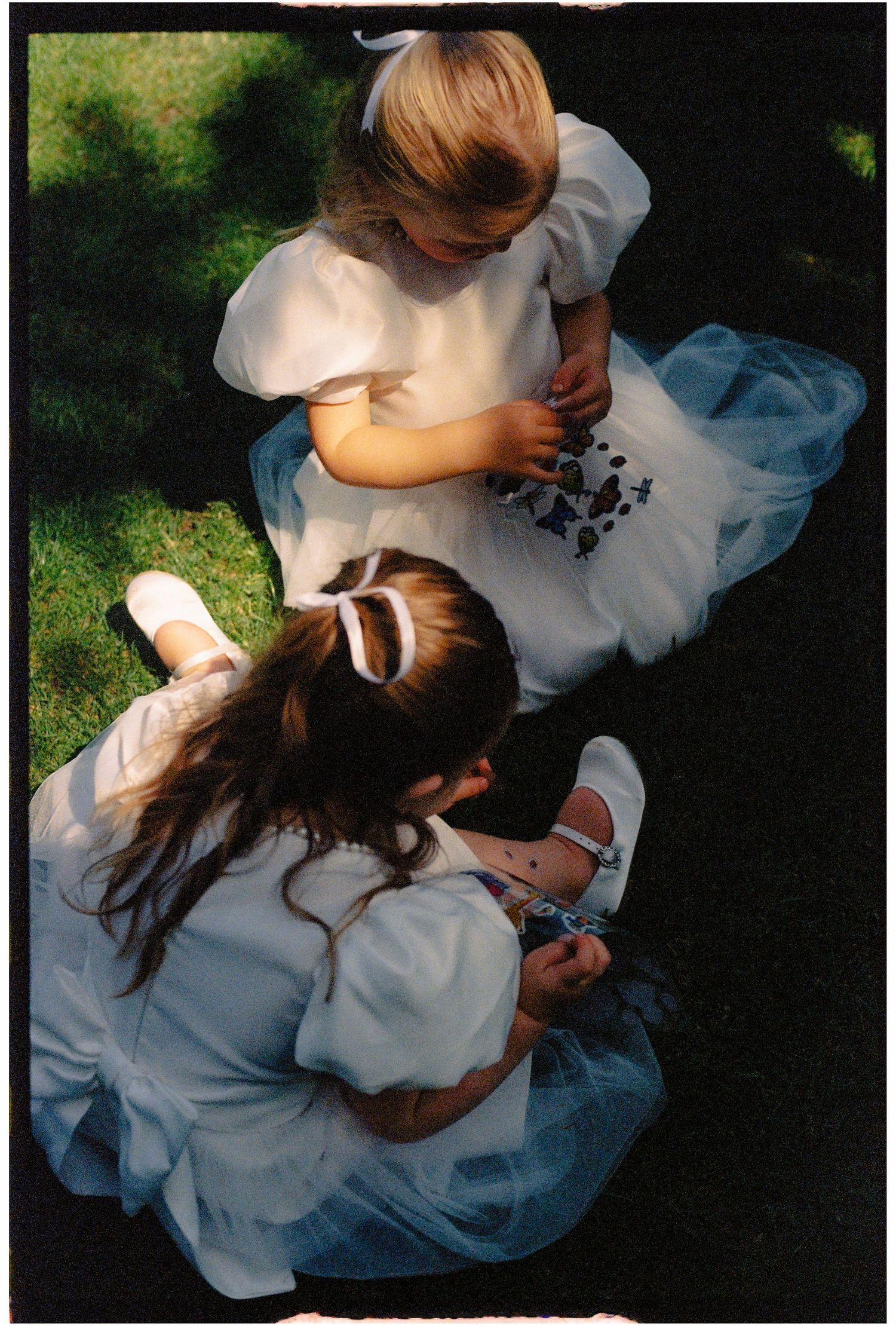 Two young flower girls in white dresses sitting on grass, examining small objects during a wedding ceremony.