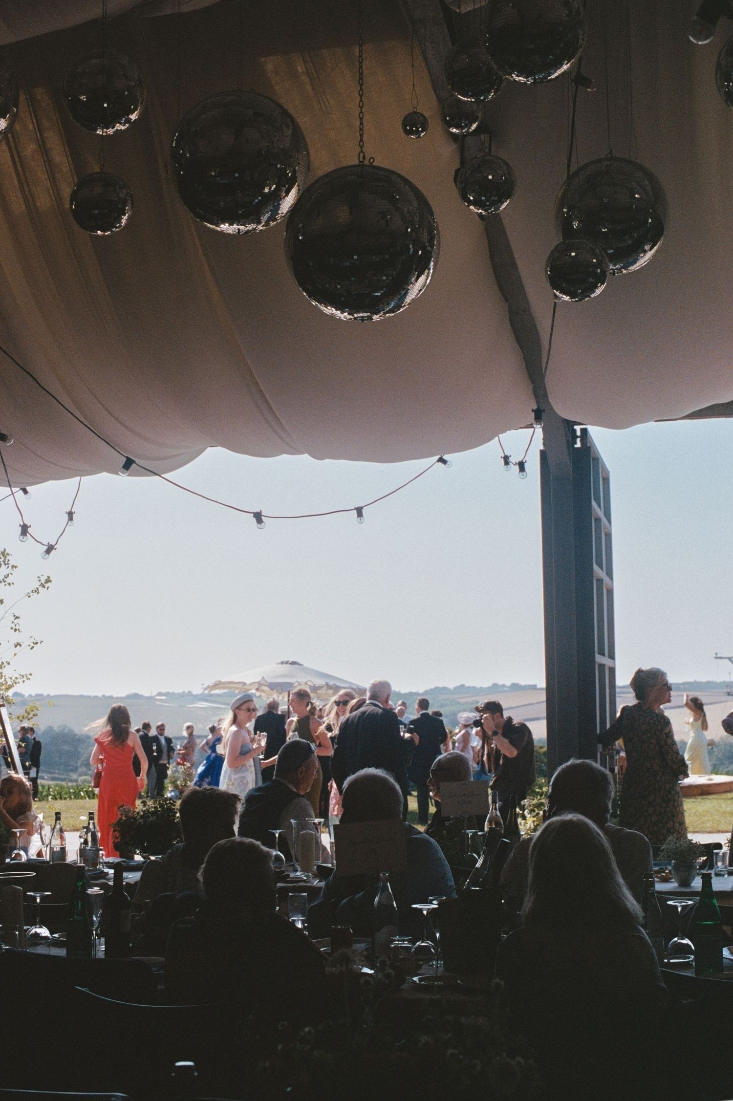 Guests mingle under a tent adorned with disco balls and string lights at an outdoor evening event.