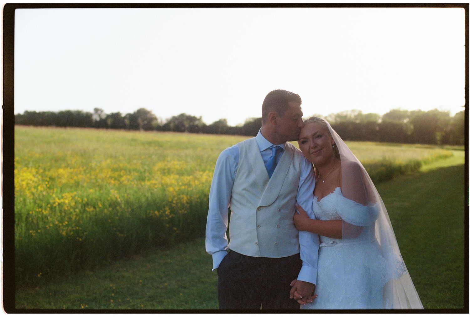 Bride and groom sharing a romantic moment in a sunlit field of yellow wildflowers on their wedding day.