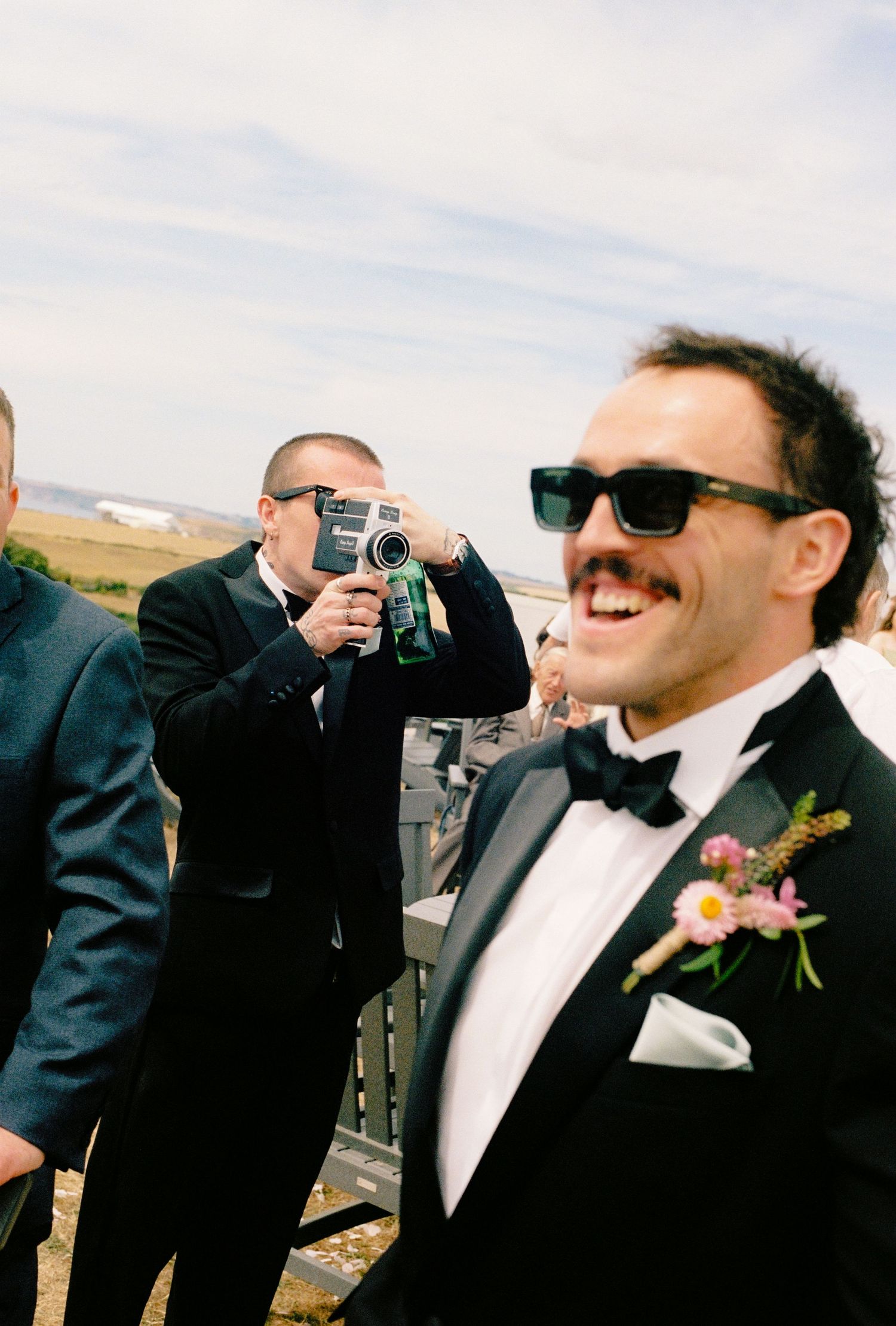 Groom in tuxedo with boutonniere smiles while guest films with vintage camera at outdoor wedding.