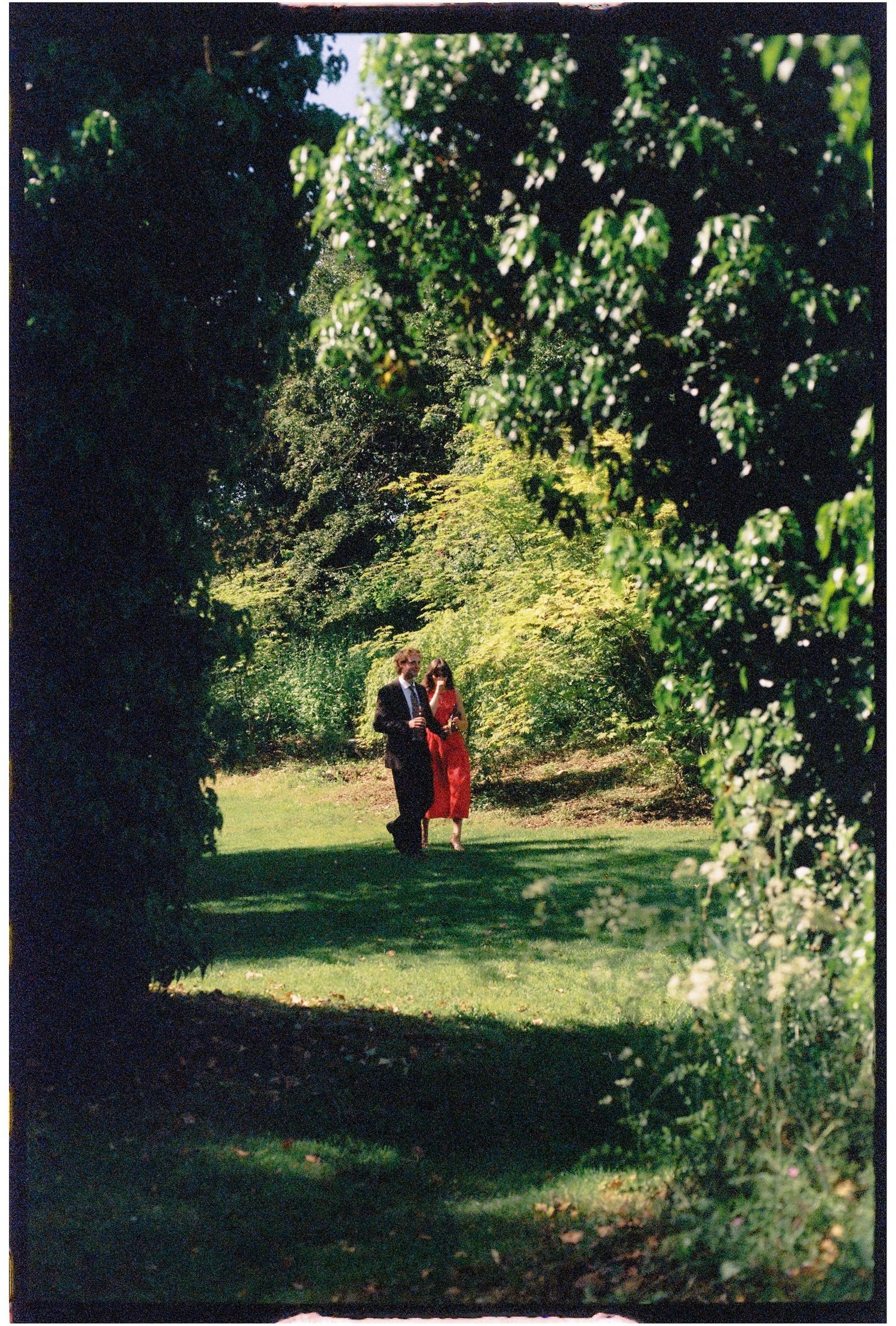 Couple in red dress and suit walking through lush green garden pathway framed by trees.