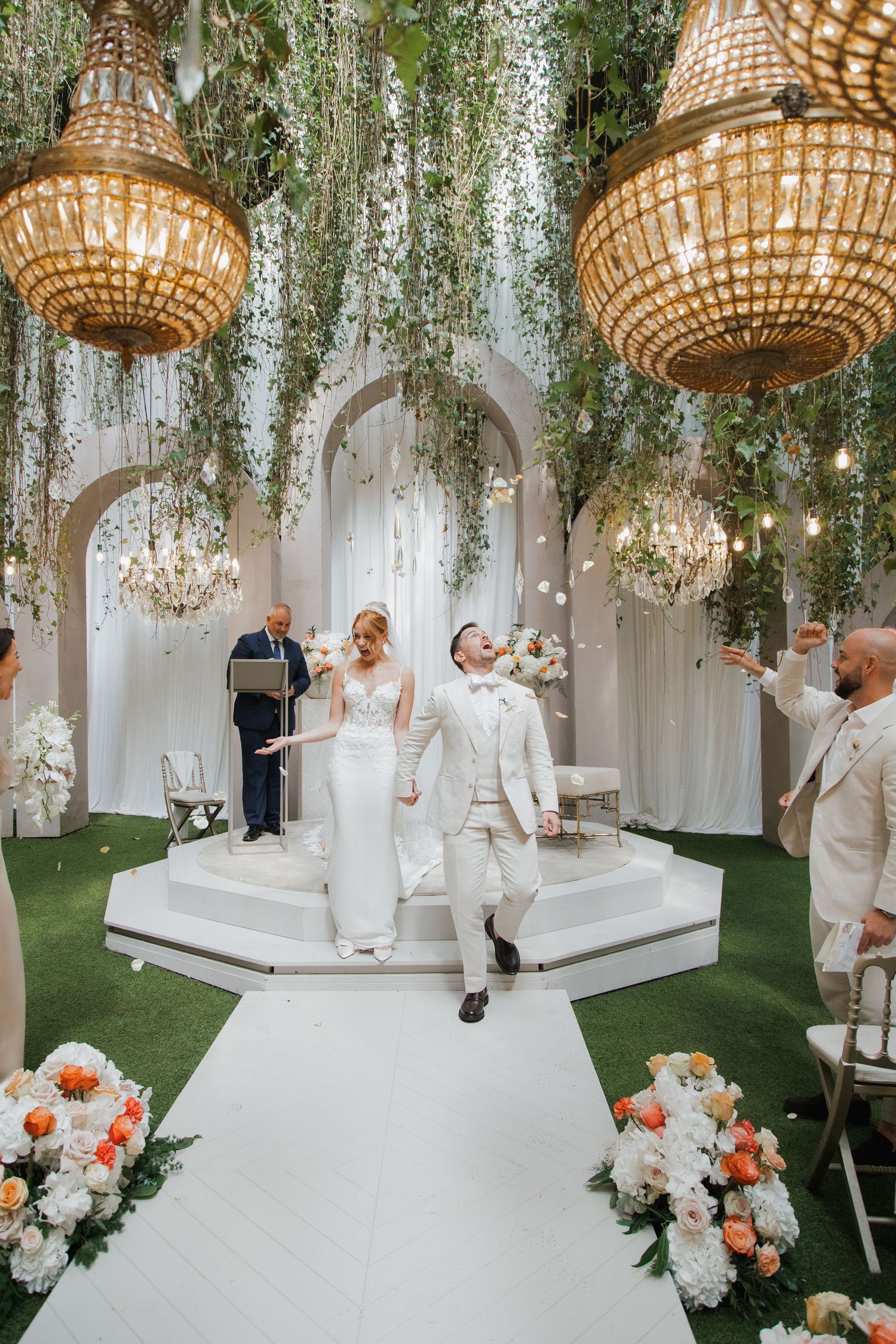 Elegant bride and groom celebrating at indoor wedding ceremony with lush greenery and grand chandeliers overhead.