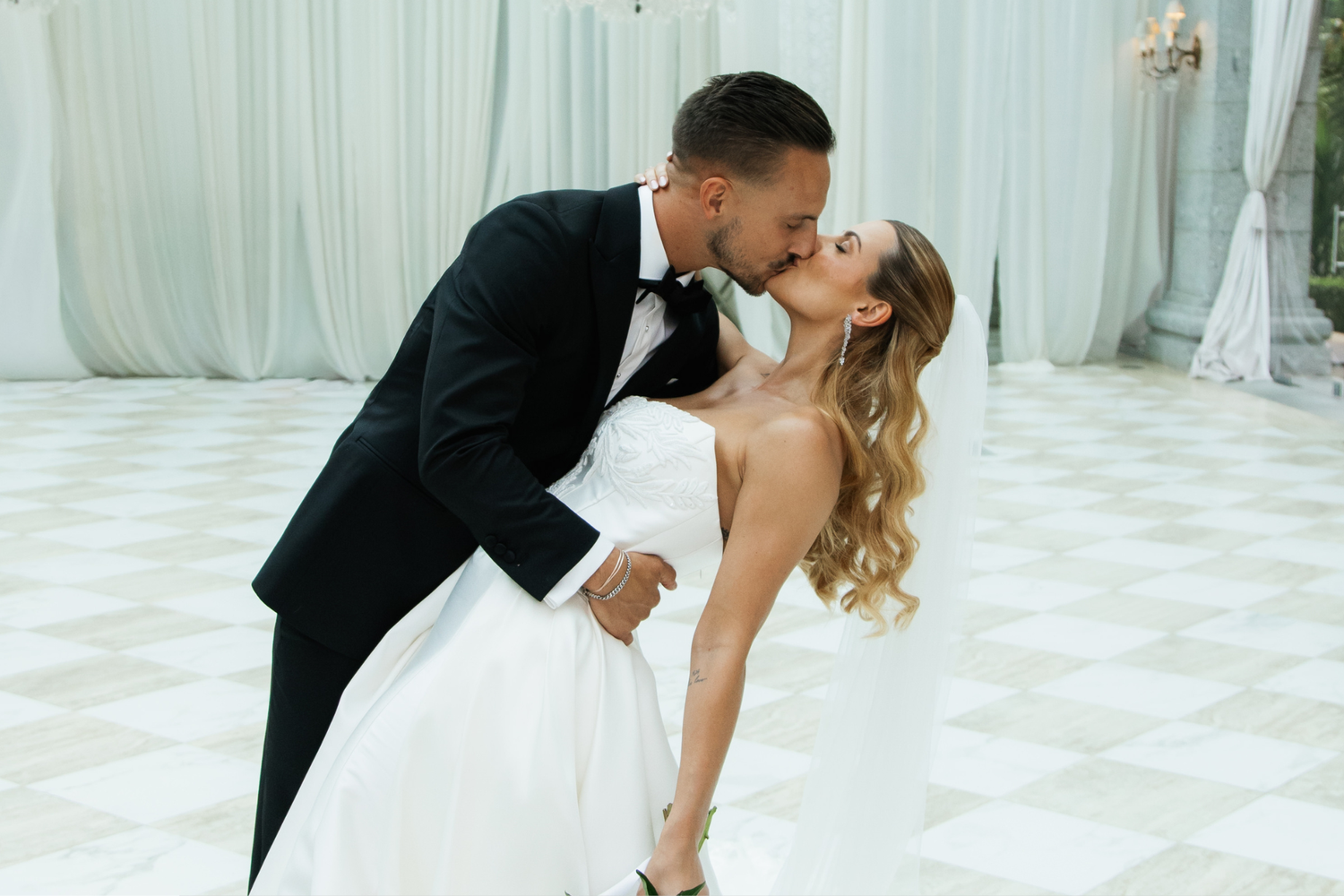 Groom passionately dips and kisses bride on elegant checkered dance floor at wedding reception.