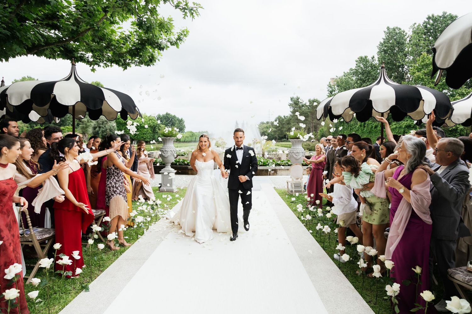 Bride and groom walk down white aisle as guests wave black-striped umbrellas during outdoor wedding ceremony.
