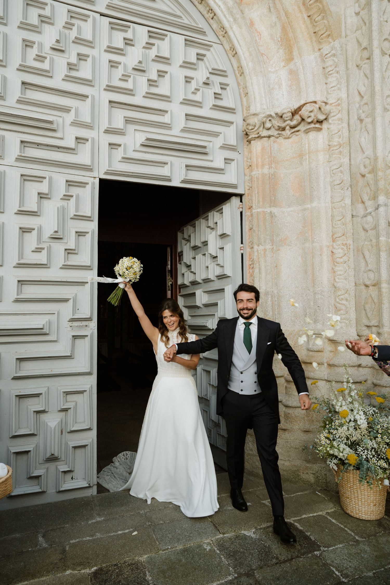 Joyful bride and groom celebrating outside ornate church doors, bride raising bouquet triumphantly.